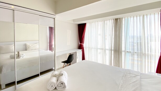 A modern hotel room with a large bed, a desk, a chair, a mirror, and a view of the city through sheer curtains.