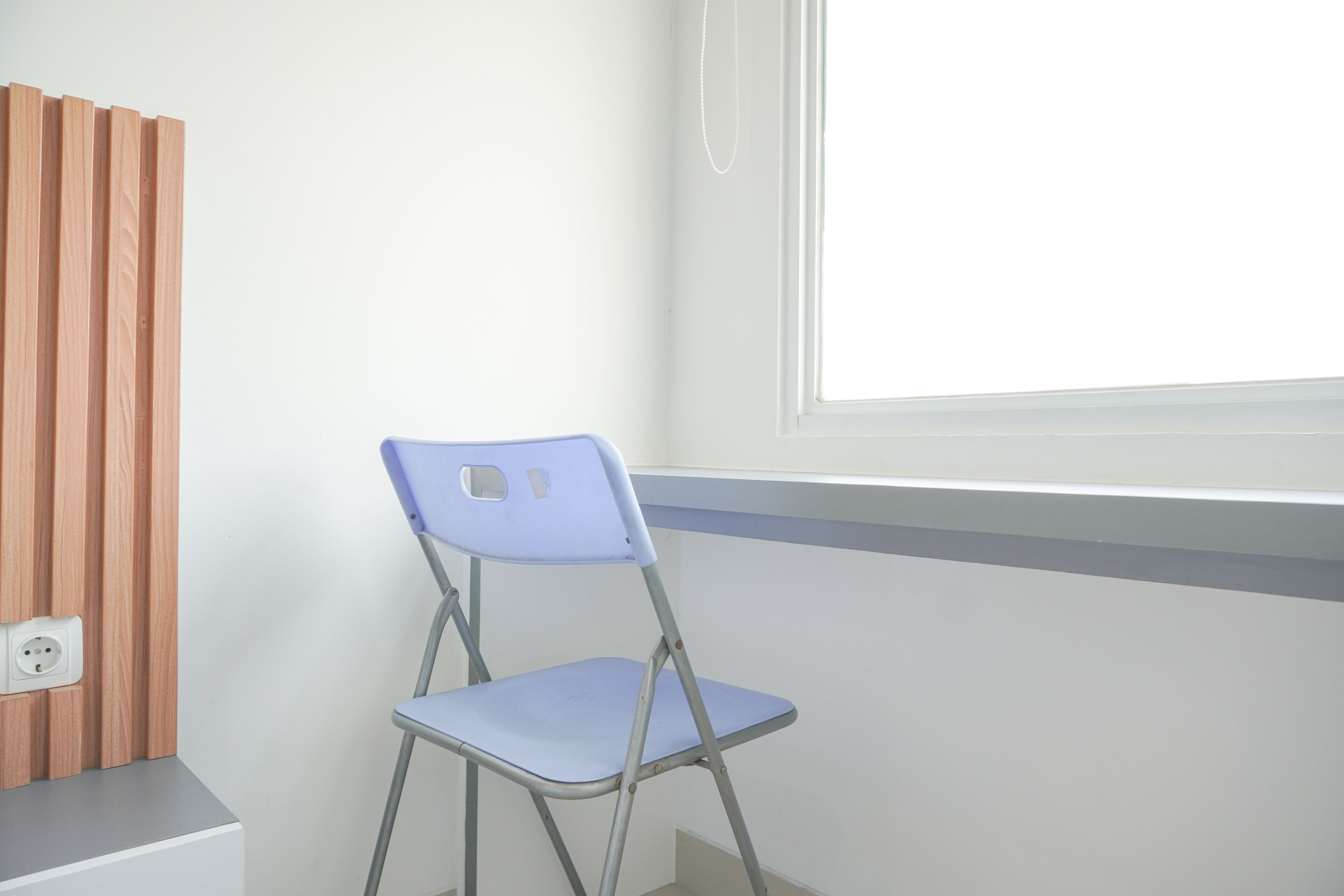 A room with a blue folding chair, a wooden panel wall, and a window with blinds.