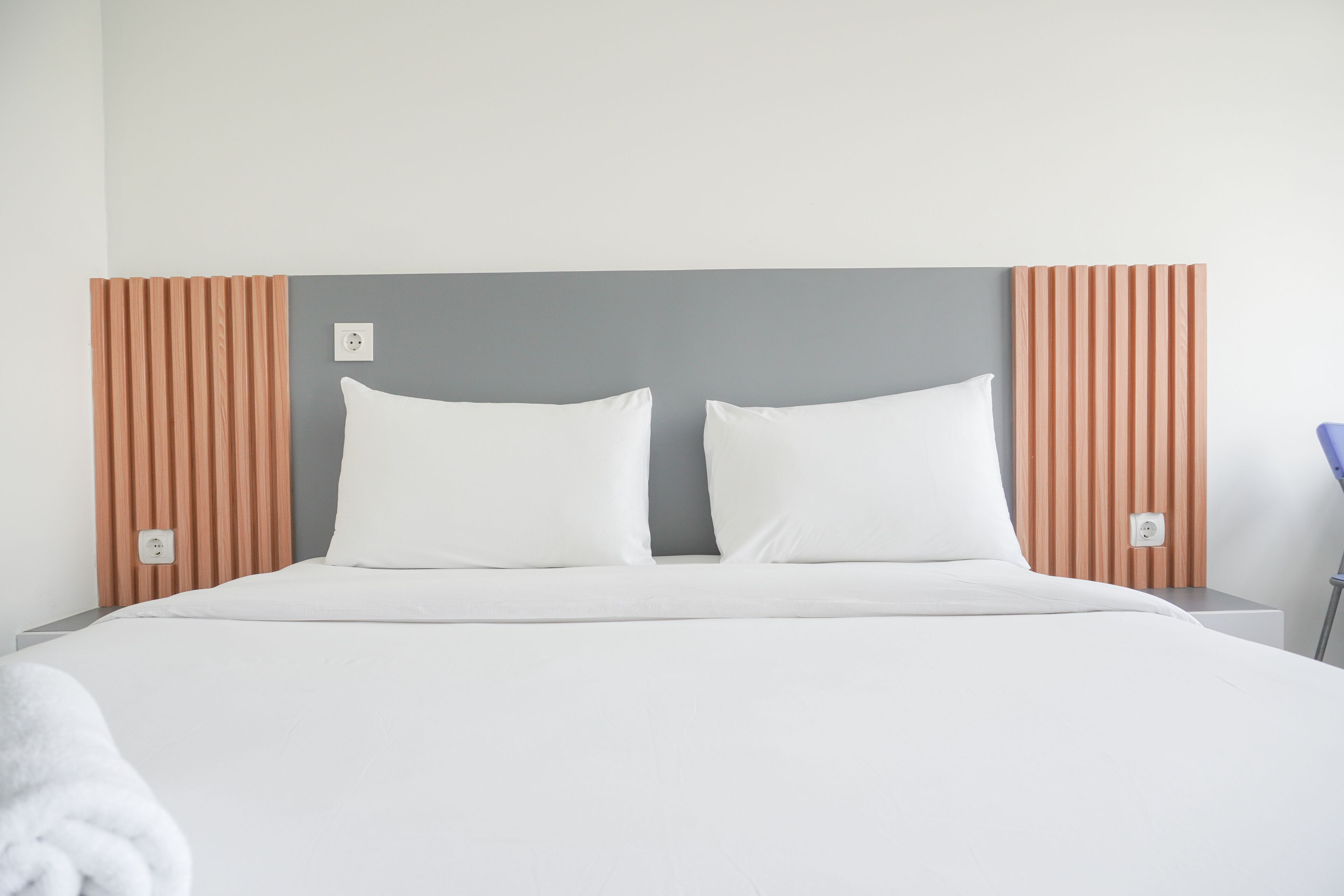 A neatly made bed with white linens and two pillows, a wooden headboard with vertical slats, and a plain white wall.