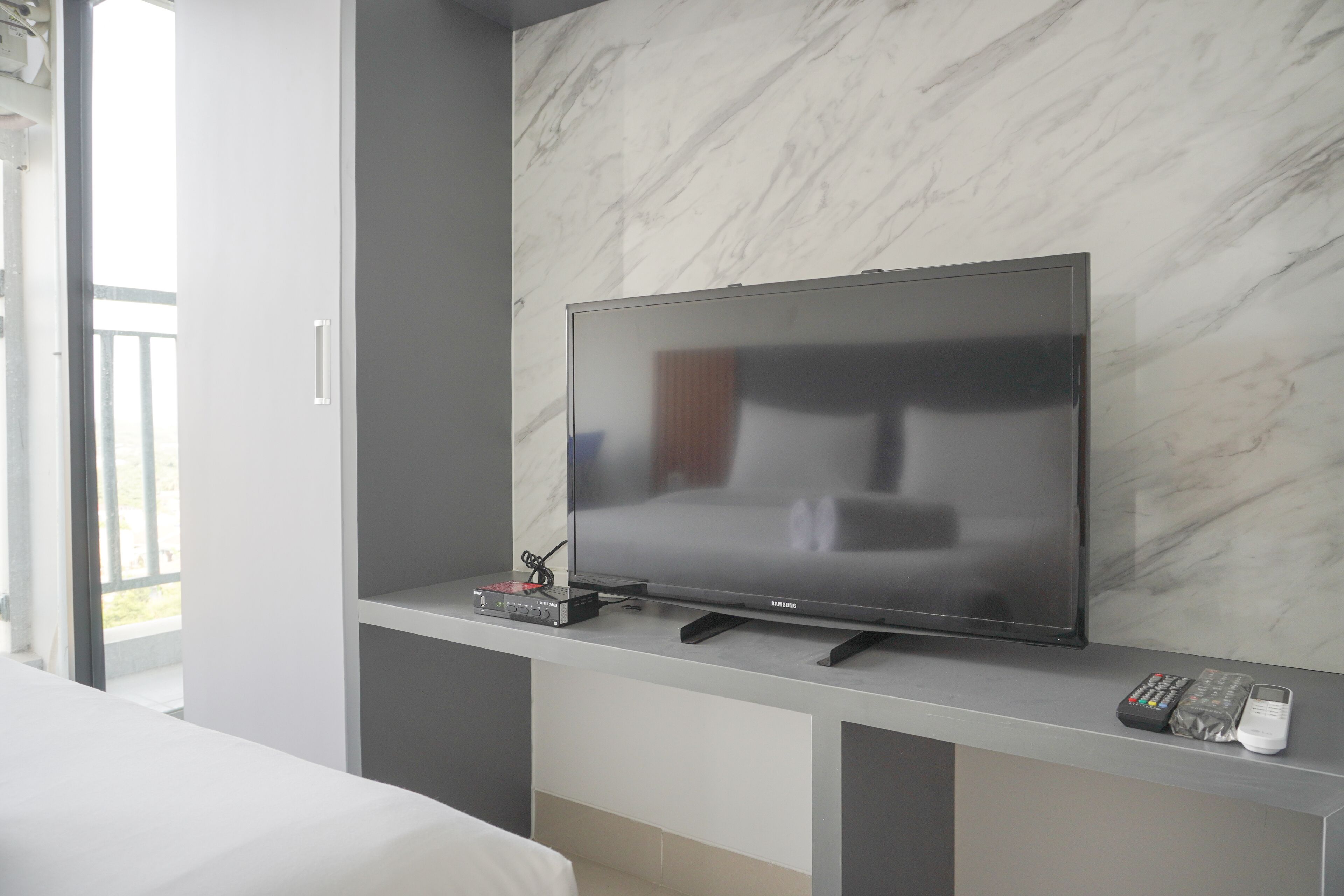A modern hotel room with a flat-screen TV mounted on a marble wall, a bedside table with a remote control, and a bed with white bedding.