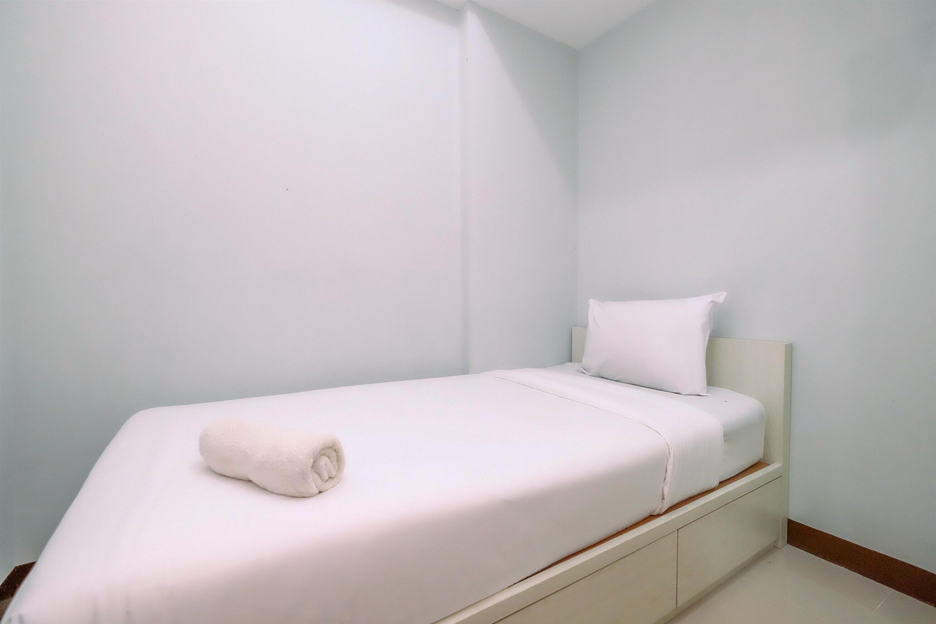 Una stanza piccola con un letto singolo, coperta bianca, un asciugamano arrotolato e un cuscino.