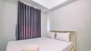 Ein ordentlich bezogenes Bett mit weißen Leinen und einem gefalteten Handtuch, ein weißer Kopfteil und ein Fenster mit dunklen Vorhängen.