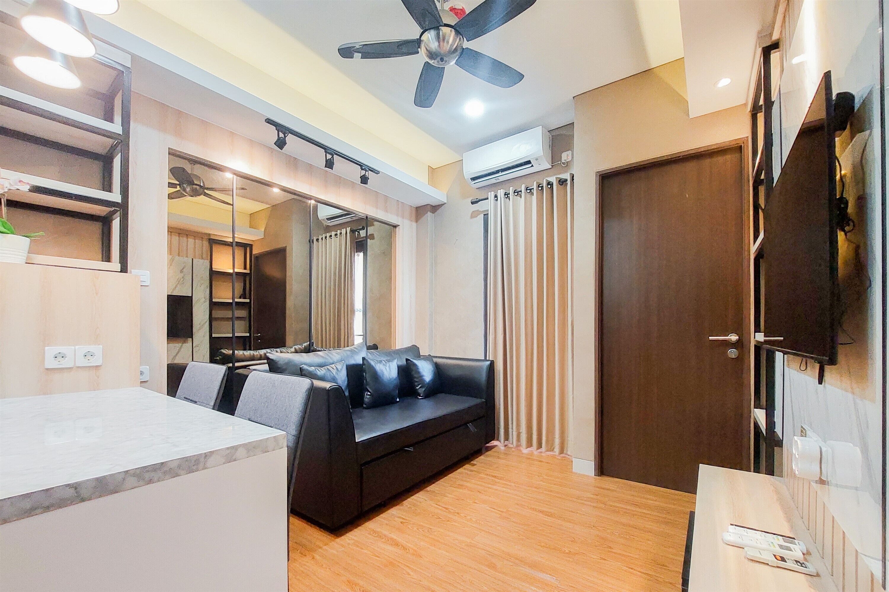 A modern living room with a black leather sofa, wooden flooring, a ceiling fan, and a TV mounted on the wall.