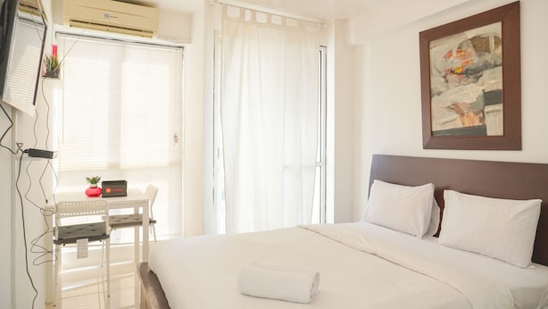 A hotel room with a bed, a desk with a chair, a window with blinds, and a painting on the wall. - Best Choice And Homey Studio Tifolia Apartment (Jakarta)