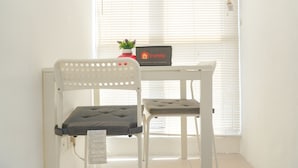 A white chair with a cushioned seat and backrest, positioned in front of a window with blinds. A small potted plant and a sign with the word "travelio" are visible on the windowsill. - Best Choice And Homey Studio Tifolia Apartment (Jakarta)