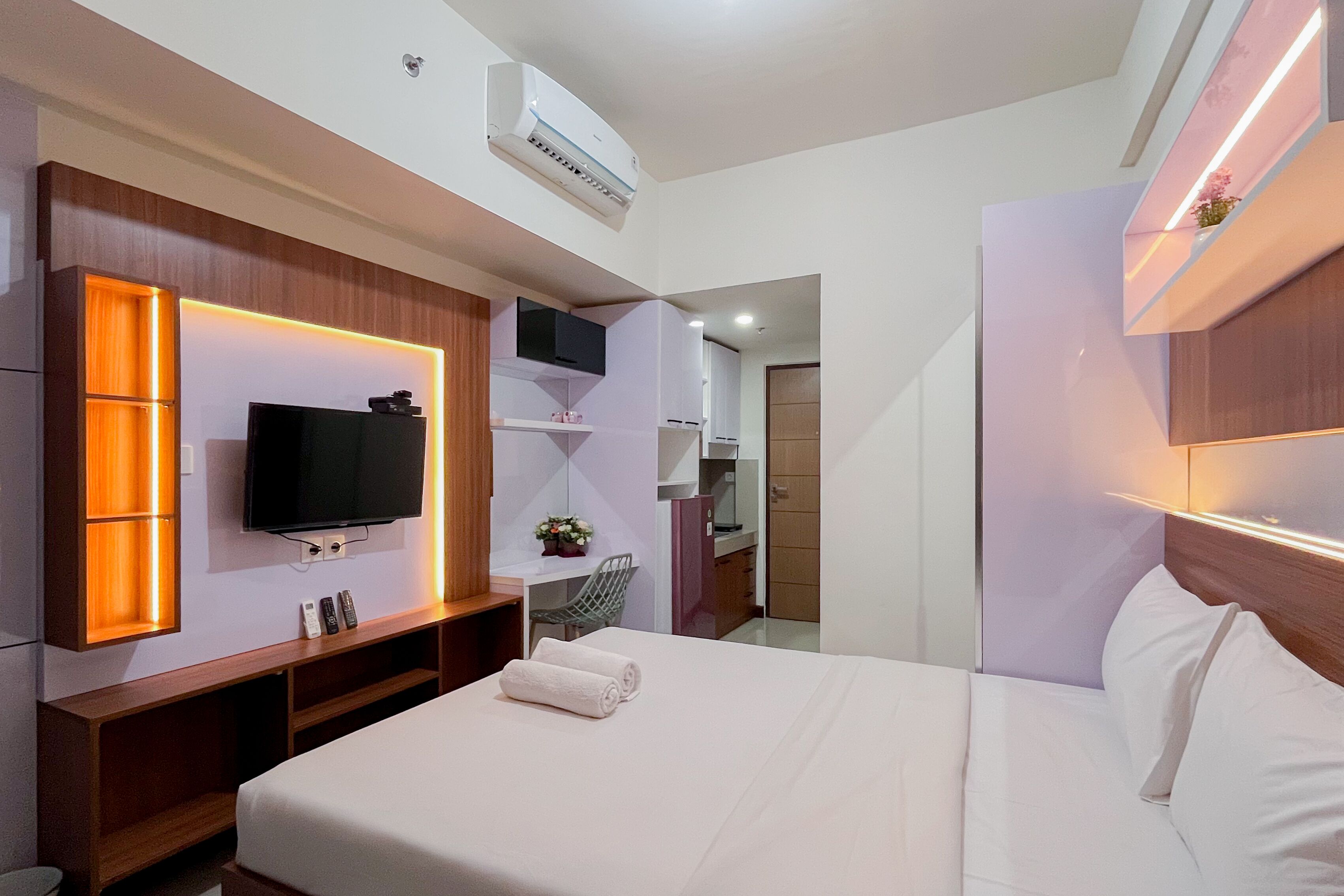 A modern hotel room with a bed, a TV mounted on the wall, a desk with a chair, and a kitchenette in the background.