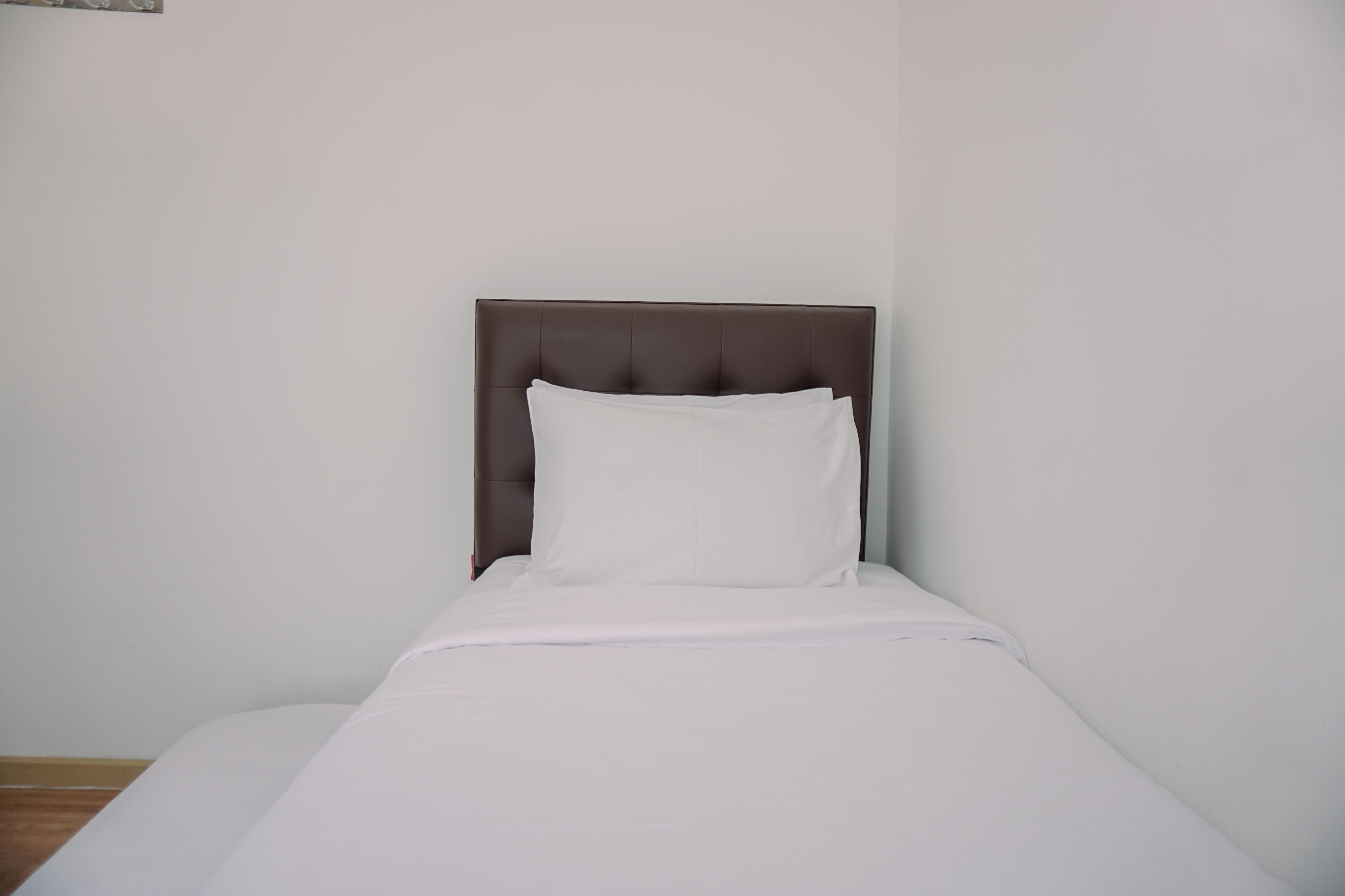 A single bed with a brown headboard and white bedding in a room with plain white walls and a wooden floor.
