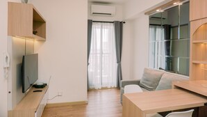 A modern, well-lit room with a wooden desk, a television, and a sofa. There is a window with curtains, an air conditioning unit, and a small shelf with a decorative item. - Modern Look 2Br Apartment M-Town Residence (Gading Serpong)