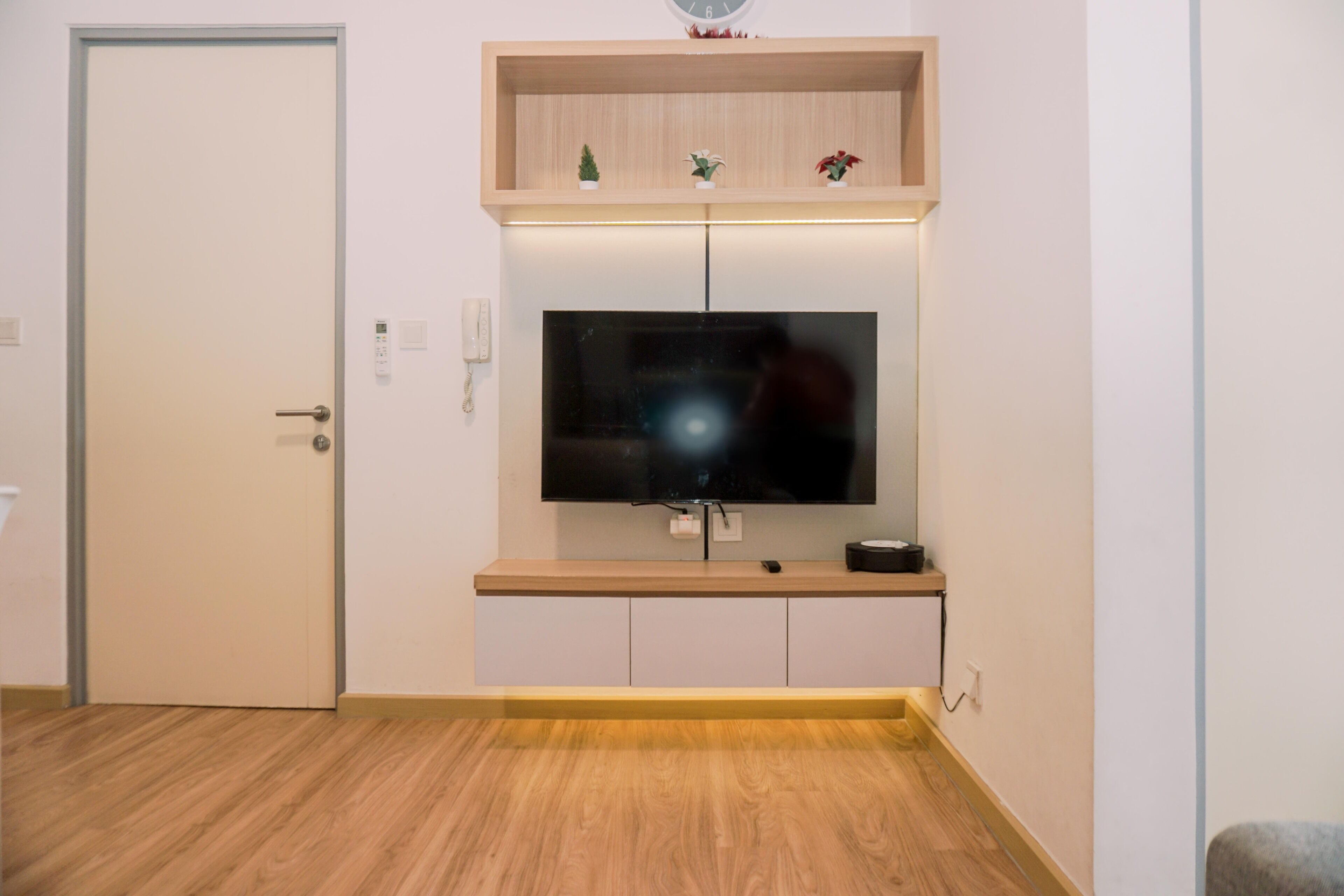Una habitación con una televisión de pantalla plana montada en un estante de madera sobre un armario blanco, una puerta a la izquierda y un suelo de color claro.