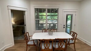 A dining area with a white table and wooden chairs, large windows with blinds, and a door leading to another room.