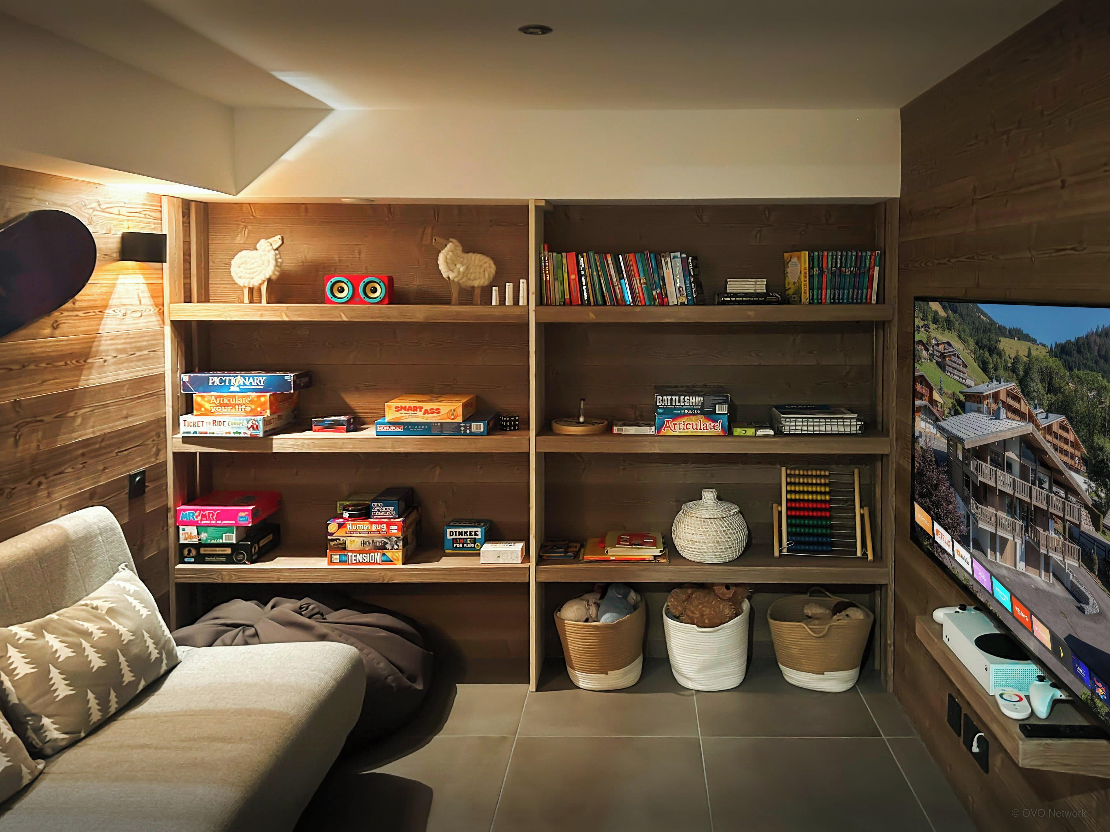 A cozy living room with a wooden bookshelf filled with board games and books, a comfortable sofa, and a television displaying a scenic view.