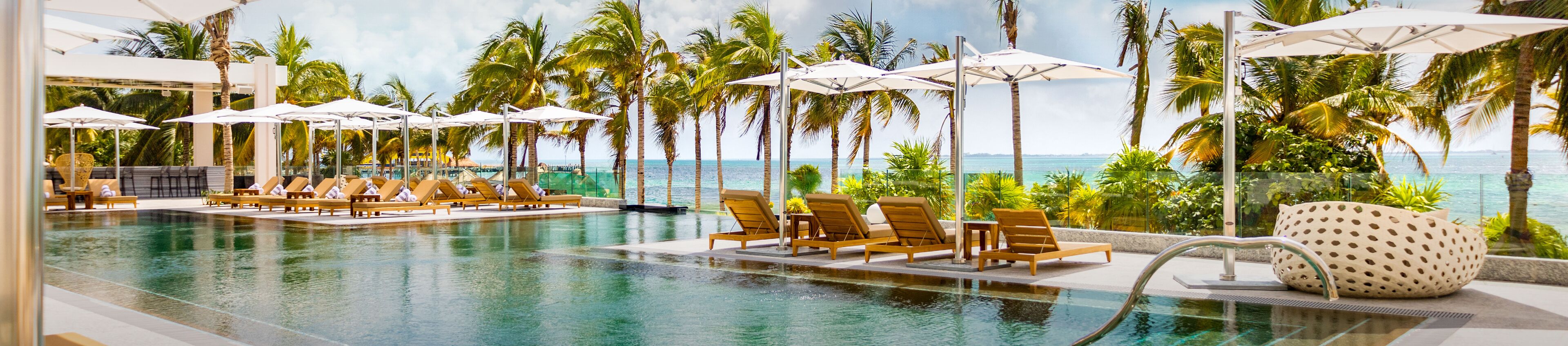 A pool area with lounge chairs and umbrellas, surrounded by palm trees and a view of the ocean.
