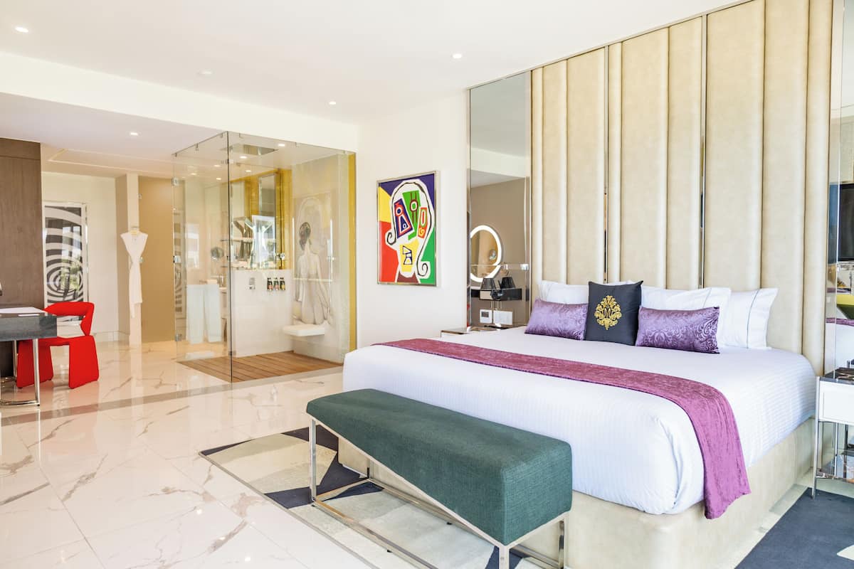 A modern hotel room with a large bed, a bench, a glass partition, a desk, and a chair.