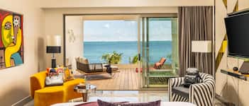 A hotel room with a bed, a yellow armchair, a striped armchair, a small table with a lamp, and a view of the sea through a sliding glass door.