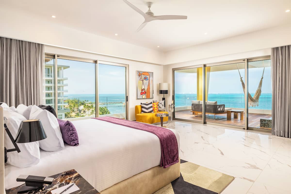 A spacious bedroom with a large bed, a ceiling fan, and a view of the ocean through floor-to-ceiling windows.