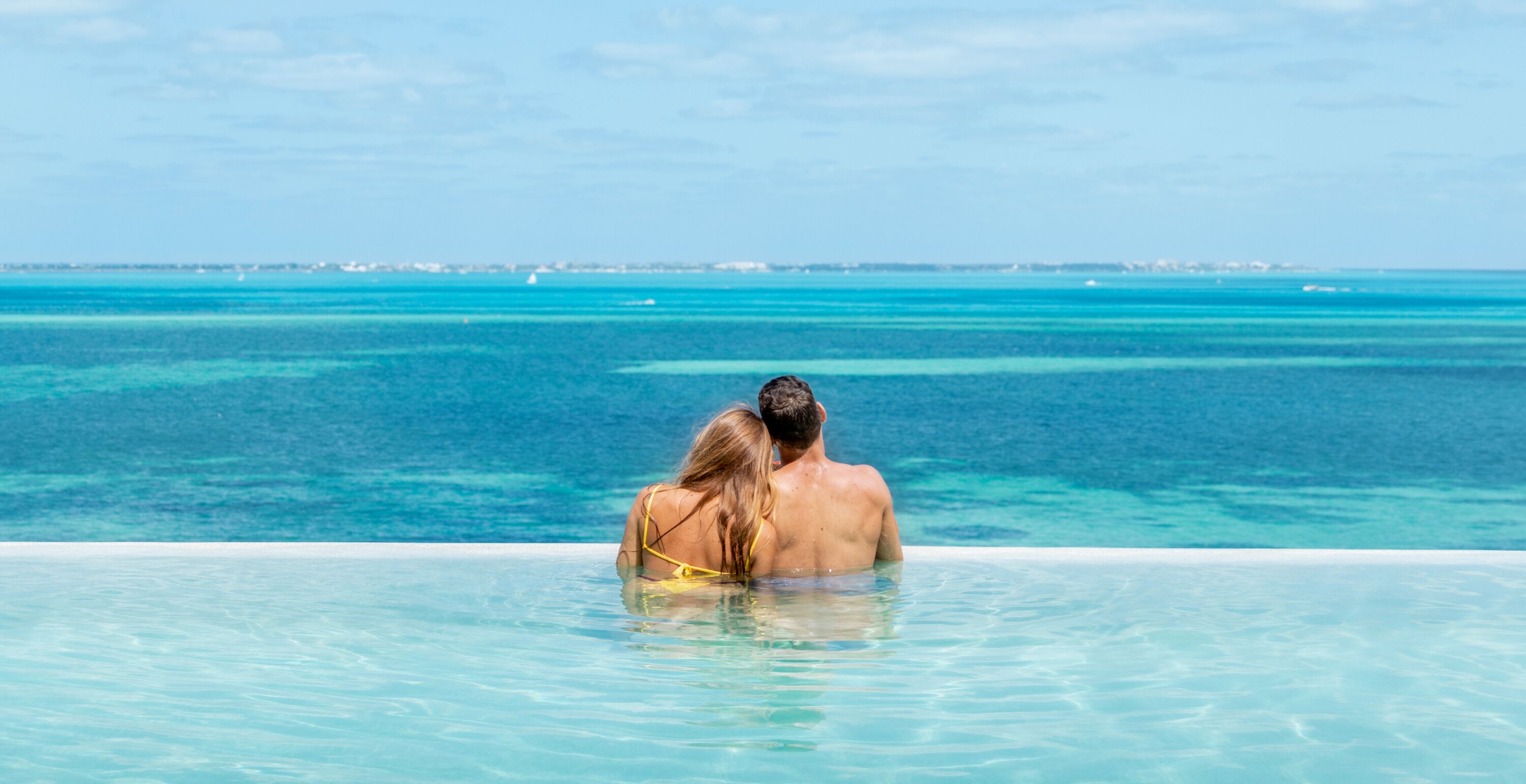 Um casal em uma piscina infinita azul turquesa, com vista para o oceano e a costa distante.