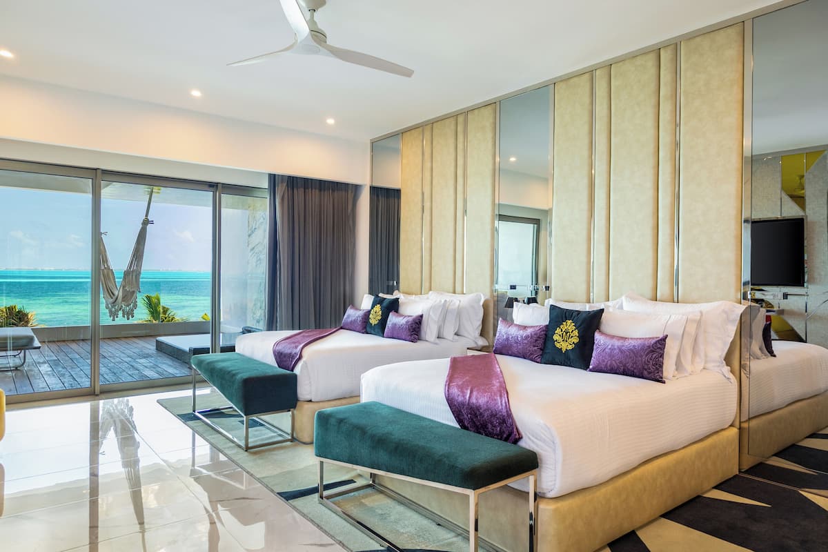A hotel room with two beds, a large window with a view of the ocean, a ceiling fan, and a flat-screen TV.