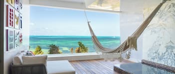 A room with a wooden floor, a hammock, a sofa, a small table, and a large window with a view of the ocean.