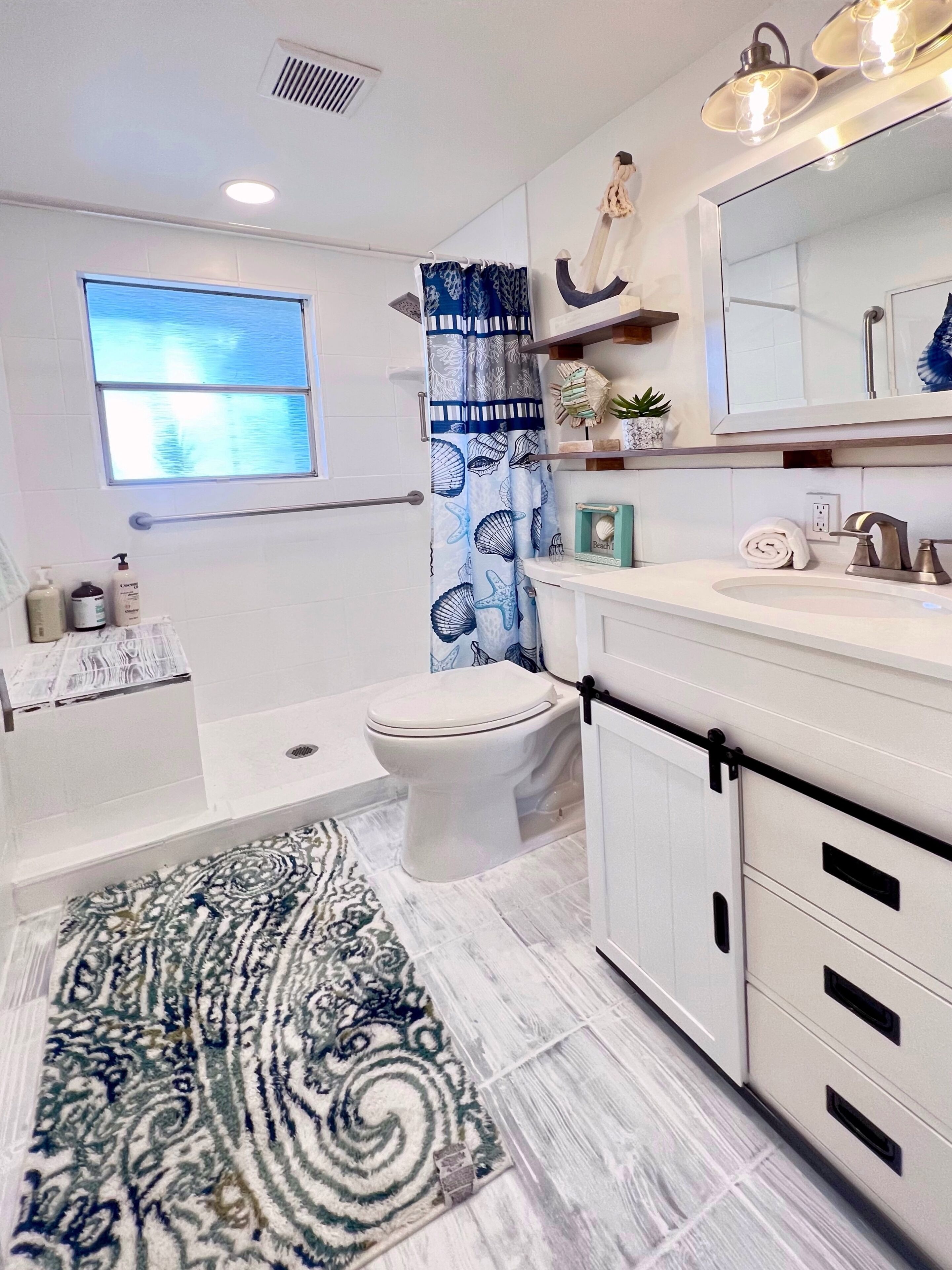A modern bathroom with a bathtub, toilet, and shower area. There is a window providing natural light, a decorative shower curtain, and a patterned rug on the floor.