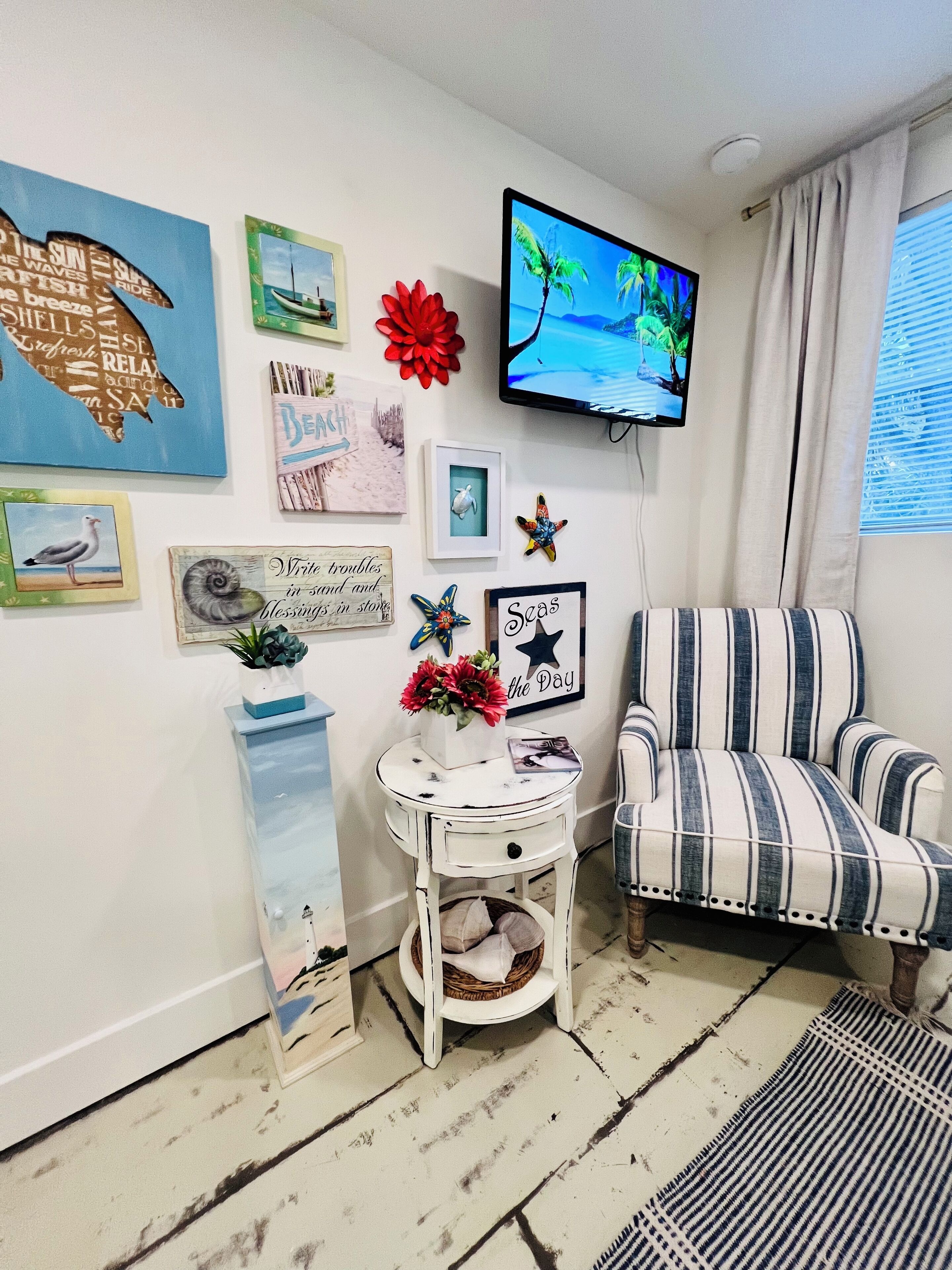 Un salón con un sillón a rayas, una mesita auxiliar con un jarrón de flores y varias obras de arte en las paredes, incluyendo una televisión que muestra una escena de playa.