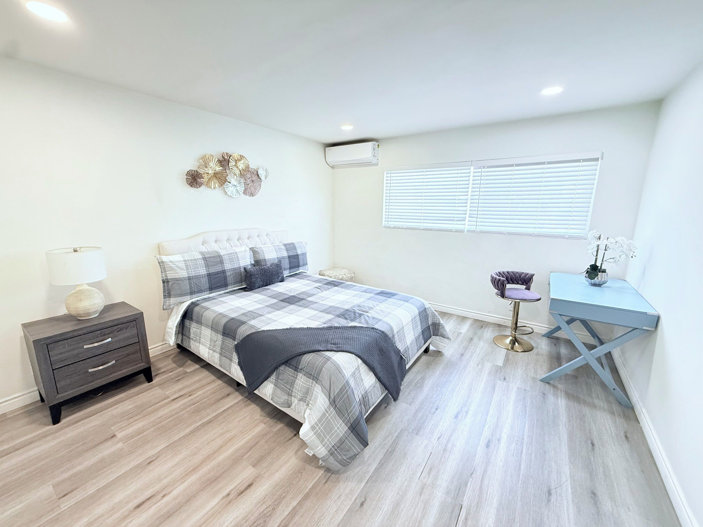 A bedroom with a bed, bedside table, lamp, decorative wall art, air conditioning unit, window with blinds, and a small desk with a chair.