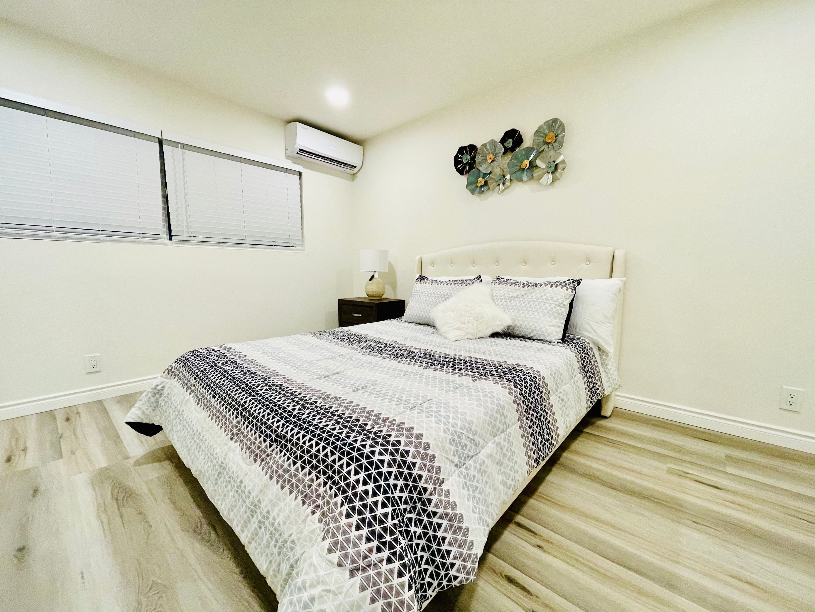 A bedroom with a bed, a nightstand, a lamp, a wall-mounted air conditioner, and a decorative wall piece.