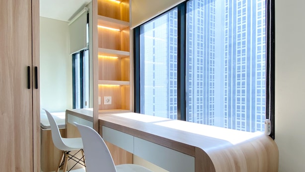 A modern bedroom with a built-in desk and seating area, large windows, and a view of tall buildings. - Comfort Stay Studio Room Pollux Chadstone Apartment (Cikarang)