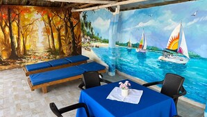A beachside restaurant with a mural of a tropical beach scene, outdoor seating with blue cushions, and a table set with a blue tablecloth and a vase of flowers. - Casa Playa James  (Cartagena)