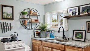 A kitchen with a stove, sink, wooden cabinets, a circular shelf with books and plants, and framed pictures on the wall. - #1 Pendleton Cottage / Midtown Hideaway / & More (Anchorage)