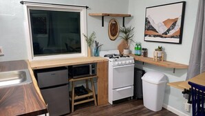 A compact kitchen with a microwave, stove, and sink. There is a small refrigerator, a trash bin, and a wooden shelf with a plant.