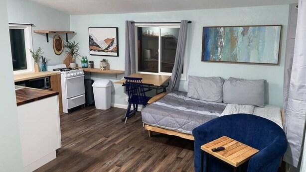 A compact room with a bed, a desk, and a chair. There is a window with curtains, a small table, and a plant.