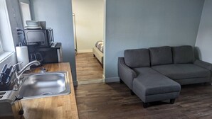 Un espace de vie compact comprenant un coin cuisine, un canapé gris et un plancher en bois.
