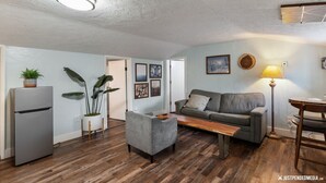 A living room with a grey sofa, a wooden coffee table, a small grey armchair, a refrigerator, and a potted plant. - Historic cottage - 10 minute walk to restaurants & trails (Anchorage)