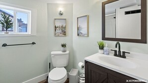 A modern bathroom with a toilet, sink, and a large mirror. There are framed pictures on the wall and a window with a view of a building outside. - Walking distance to restaurants, trails, and cafes.  10 min. uber from airport. (Anchorage)