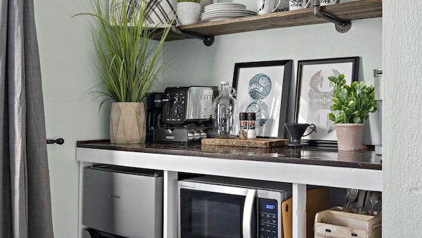 A compact kitchen with a microwave, coffee maker, and a small refrigerator. There are potted plants and a shelf with cups and a coffee pot. - 1964 Earthquake Cottage At Slumber Village (Anchorage)
