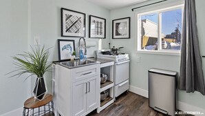 A compact kitchen with white cabinets, a sink, a stove, and a trash can. There are framed pictures on the wall and a plant on a small table. - 1964 Earthquake Cottage At Slumber Village (Anchorage)