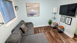 A living room with a grey sofa, a flat-screen TV mounted on the wall, a wooden floor, and a window with blinds. - 1964 Earthquake Cottage At Slumber Village (Anchorage)