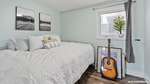 A bedroom with a bed, two framed pictures, a guitar, a window with a plant, and a radiator. - 1964 Earthquake Cottage At Slumber Village (Anchorage)