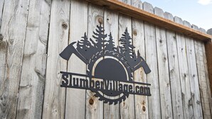 Una cerca de madera con un logo que incluye una hoja de sierra, un martillo y un árbol, y el texto "SlumberVillage.com".