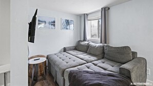 Una habitación con un sofá gris, una pequeña mesa redonda de madera y una ventana con cortinas.