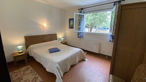 A bedroom with a wooden headboard, a bed with a white bedspread, a nightstand with a lamp, a window with curtains, and a radiator. - 085 - Invitation to rest in this villa in Grande Combe (SOULAC SUR MER)