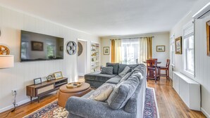 Uma sala de estar com um sofá cinza grande, uma televisão de tela plana, uma mesa de centro de madeira e um tapete estampado.