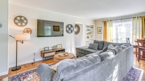 Uma sala de estar com um sofá modular cinza, uma TV de tela plana na parede, um abajur e uma pequena mesa lateral de madeira com objetos decorativos.