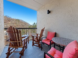 A balcony with wooden chairs and a table, a view of a hillside, and a textured wall.
