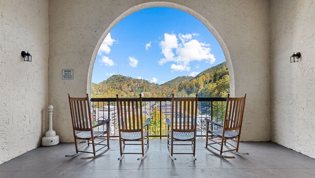 Ein Balkon mit vier hölzernen Schaukelstühlen, der einen idyllischen Blick auf eine Berglandschaft und eine Stadt bietet.