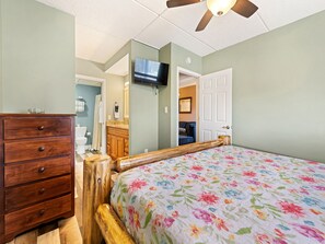 A bedroom with a wooden bed frame, a dresser, a television mounted on the wall, and a door leading to another room.
