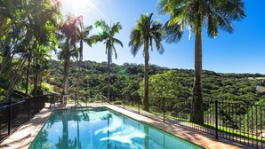 Une piscine avec une terrasse en bois, entourée d’une clôture noire, et des palmiers en arrière-plan.
