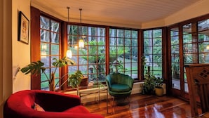 Une pièce dotée de grandes fenêtres, d’un fauteuil rouge, d’un fauteuil vert et de plusieurs plantes en pot.