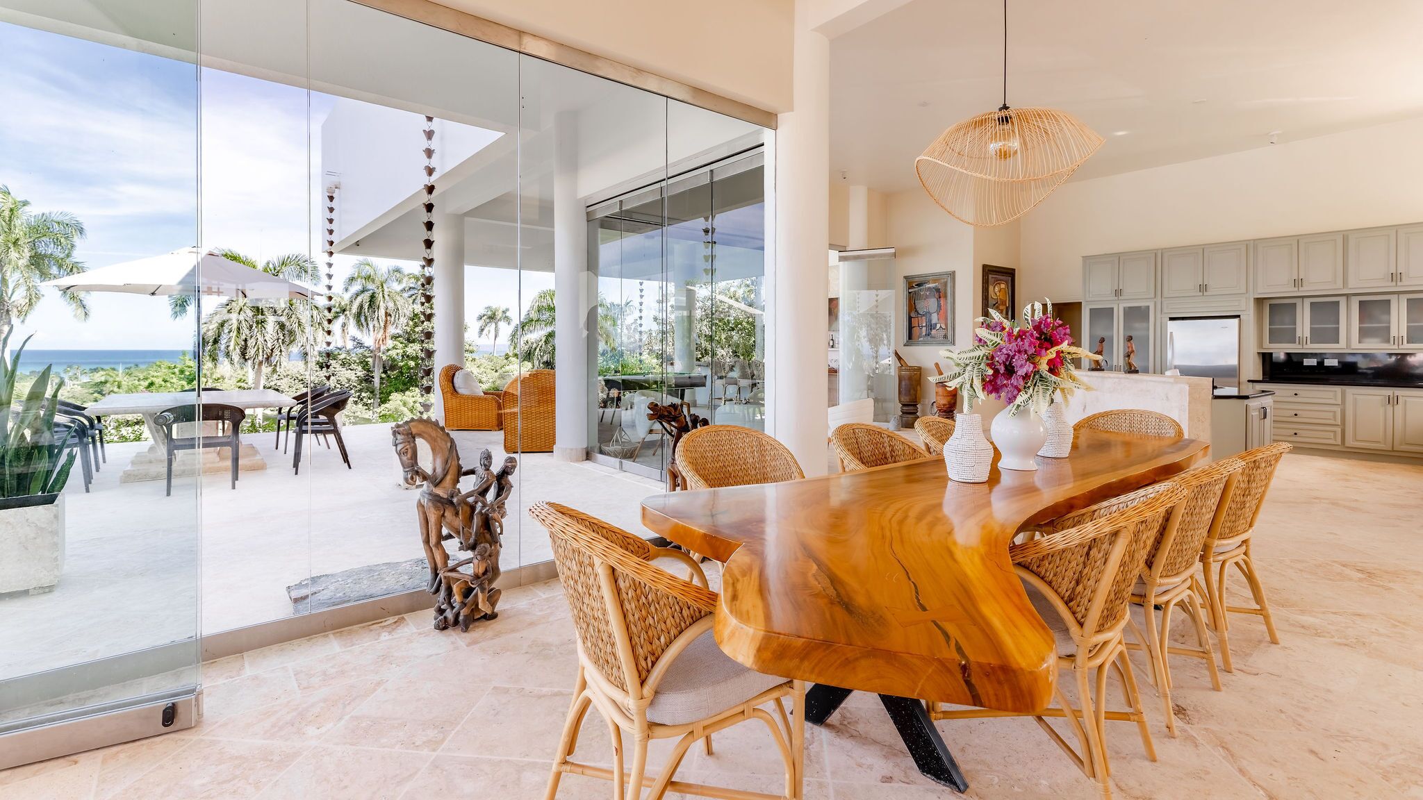 Un vaste espace dédié à la salle à manger, avec une table en bois et des chaises en osier, donnant sur un jardin tropical et l’océan.