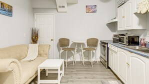 Cocina compacta con armarios blancos, microondas y un pequeño comedor con mesa blanca y sillas de mimbre.